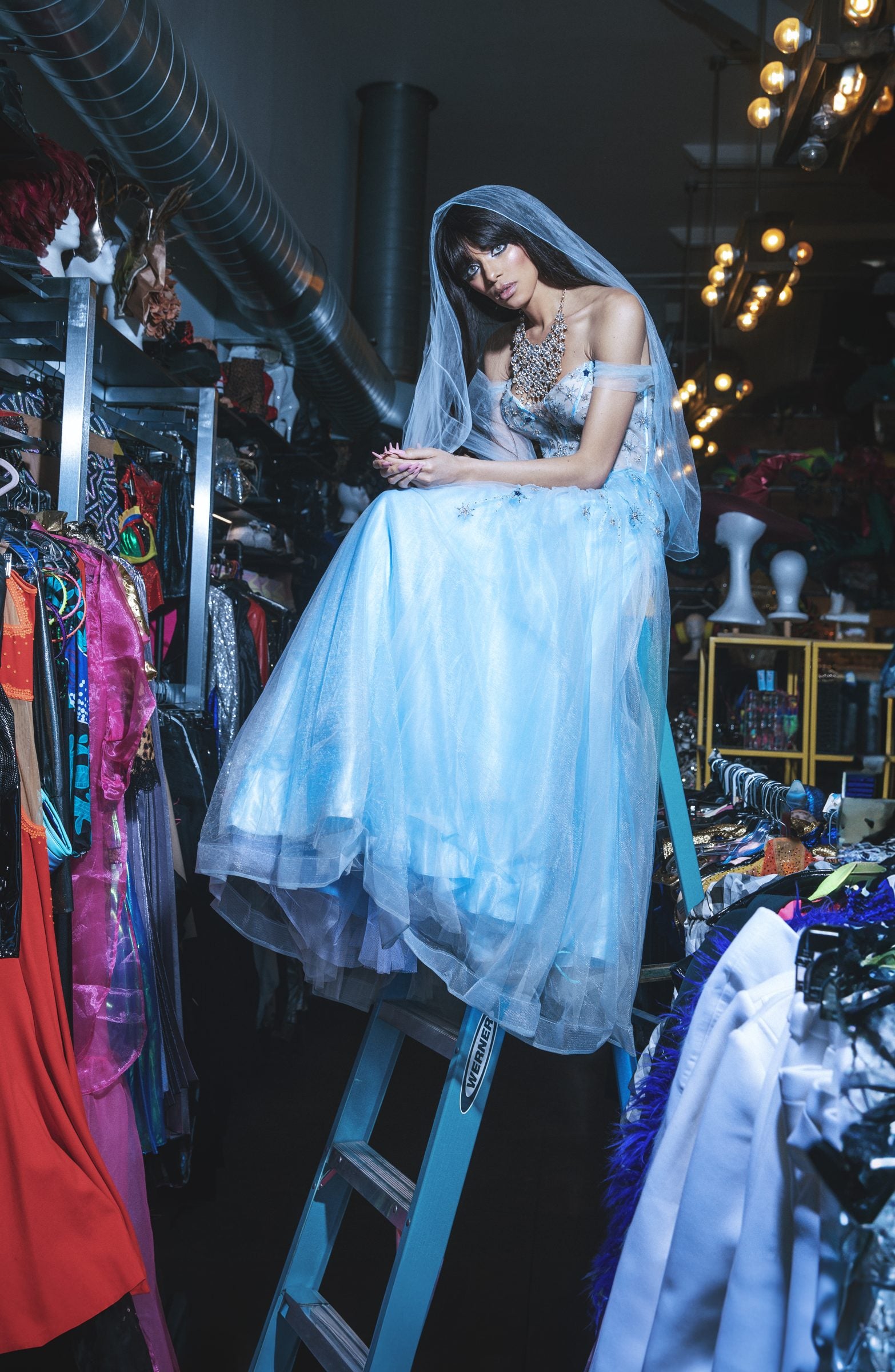 A model in a pale blue tulle gown and veil seated on a ladder among the archive rails